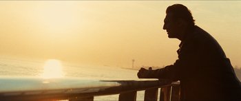 Movie still from “Taken 2” (2012), directed by Olivier Megaton – A man sitting on top of a wooden railing near the ocean; Medium shot, Low angle