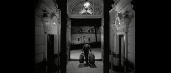 Movie still from “Talk to Her” (2002), directed by Pedro Almodóvar – A man holding two suitcases standing in an archway; Wide shot, Low angle
