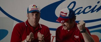Movie still from “Talladega Nights: The Ballad of Ricky Bobby” (2006), directed by Adam McKay – Two men in red and white hats are talking to each other in front of a white background; Close Up shot, Low angle