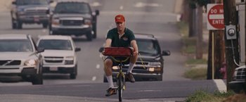 Movie still from “Talladega Nights: The Ballad of Ricky Bobby” (2006), directed by Adam McKay – A man riding a bike down a street with cars behind him; Wide shot, High angle