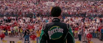 Movie still from “Talladega Nights: The Ballad of Ricky Bobby” (2006), directed by Adam McKay – A man in a racing suit standing in front of a crowd of people; Wide shot, Over the shoulder angle