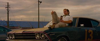 Movie still from “Talladega Nights: The Ballad of Ricky Bobby” (2006), directed by Adam McKay – A man sitting on the hood of a car while holding a cell phone; Medium shot, Low angle