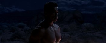 Movie still from “Terminator 3: Rise of the Machines” (2003), directed by Jonathan Mostow – A man with no shirt in the dark with mountains in the background; Medium shot, Low angle