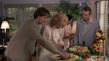 Movie still from “Terms of Endearment” (1983), directed by James L. Brooks – A group of people standing around a table with food on top of it; Medium shot, High angle