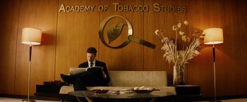 Movie still from “Thank You for Smoking” (2005), directed by Jason Reitman – A man sitting at a table in front of a sign that reads " academy of tobacco studies "; Wide shot, Low angle
