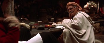 Movie still from “The 13th Warrior” (1999), directed by Michael Crichton – An old man sitting at a table with a bowl of fruit; Medium shot, Over the shoulder angle