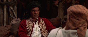 Movie still from “The 13th Warrior” (1999), directed by Michael Crichton – A man in a red and white outfit sitting on a couch; Close Up shot, Over the shoulder angle