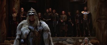 Movie still from “The 13th Warrior” (1999), directed by Michael Crichton – A group of people standing in a room with a man in a costume; Medium shot, Over the shoulder angle