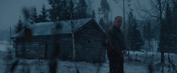 Movie still from “Out Stealing Horses” (2019), directed by Hans Petter Moland – A man standing in front of a log cabin in the snow; Wide shot, Low angle