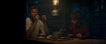 Movie still from “Out Stealing Horses” (2019), directed by Hans Petter Moland – Two men sitting at a wooden table eating food; Medium shot, Low angle