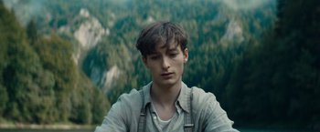 Movie still from “Out Stealing Horses” (2019), directed by Hans Petter Moland – A young man in a gray shirt is standing in front of a forest; Close Up shot, High angle