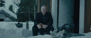 Movie still from “Out Stealing Horses” (2019), directed by Hans Petter Moland – An older man sitting on the ground in the snow; Medium shot, Over the shoulder angle