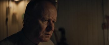 Movie still from “Out Stealing Horses” (2019), directed by Hans Petter Moland – An older man looking at the camera in a dimly - lit room; Close Up shot, Over the shoulder angle