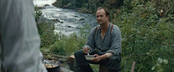 Movie still from “Out Stealing Horses” (2019), directed by Hans Petter Moland – A man sitting on a log near a river; Medium shot, Over the shoulder angle