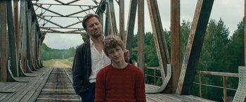 Movie still from “Out Stealing Horses” (2019), directed by Hans Petter Moland – A man standing next to a young boy on a bridge; Medium shot, Low angle