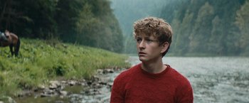 Movie still from “Out Stealing Horses” (2019), directed by Hans Petter Moland – A young man wearing a red sweater standing next to a body of water; Medium shot, Over the shoulder angle