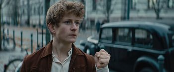 Movie still from “Out Stealing Horses” (2019), directed by Hans Petter Moland – A young man in a brown jacket is standing in front of a black car; Close Up shot, Low angle