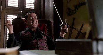 Movie still from “The Addams Family” (1991), directed by Barry Sonnenfeld – A man sitting in front of a laptop holding a sword; Medium shot, Low angle