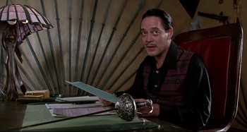 Movie still from “The Addams Family” (1991), directed by Barry Sonnenfeld – A man sitting at a table holding a sword; Medium shot, Low angle