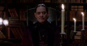 Movie still from “The Addams Family” (1991), directed by Barry Sonnenfeld – A man sitting in a chair in front of a lit candle; Medium shot, Low angle