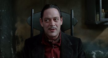 Movie still from “The Addams Family” (1991), directed by Barry Sonnenfeld – A man sitting in a chair wearing a red shirt and black jacket; Close Up shot, Low angle