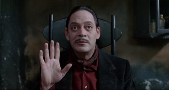 Movie still from “The Addams Family” (1991), directed by Barry Sonnenfeld – A man sitting in a chair with his hand raised; Close Up shot, Over the shoulder angle