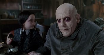 Movie still from “The Addams Family” (1991), directed by Barry Sonnenfeld – A man with a bald head and black eyes sitting in front of a girl; Close Up shot, Over the shoulder angle