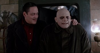 Movie still from “The Addams Family” (1991), directed by Barry Sonnenfeld – A man with a bald head is standing next to another man in the back of a car; Medium shot, Low angle