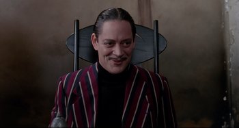 Movie still from “The Addams Family” (1991), directed by Barry Sonnenfeld – A man sitting in a chair wearing a striped jacket; Medium shot, Low angle
