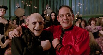 Movie still from “The Addams Family” (1991), directed by Barry Sonnenfeld – Two people posing for a picture with a mask on; Medium shot, Low angle