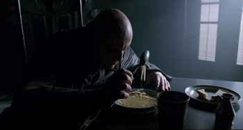 Movie still from “The Addams Family” (1991), directed by Barry Sonnenfeld – A bald man sitting at a table with a bowl of noodles; Medium shot, Low angle
