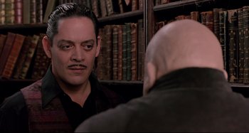 Movie still from “The Addams Family” (1991), directed by Barry Sonnenfeld – A man with a mustache is talking to another man in front of a book shelf; Close Up shot, Over the shoulder angle