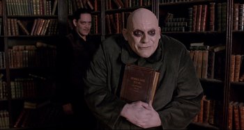 Movie still from “The Addams Family” (1991), directed by Barry Sonnenfeld – A man with a bald head holding an old book; Medium shot, Low angle