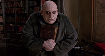 Movie still from “The Addams Family” (1991), directed by Barry Sonnenfeld – A man with a bald head holding a book; Medium shot, Low angle