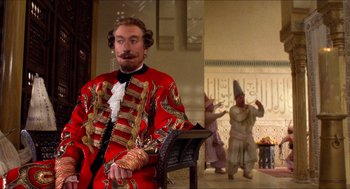 Movie still from “The Adventures of Baron Munchausen” (1988), directed by Terry Gilliam – A man in a red and gold suit sitting on a chair; Medium shot, Over the shoulder angle