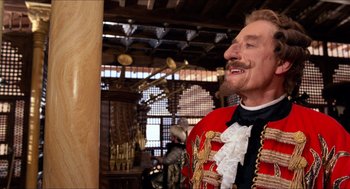Movie still from “The Adventures of Baron Munchausen” (1988), directed by Terry Gilliam – A man with a beard and a red jacket is standing in front of a wooden structure; Medium shot, Over the shoulder angle