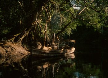 Movie still from “The African Queen” (1951), directed by John Huston – A boat floating on a body of water next to trees; Extreme Wide shot, High angle