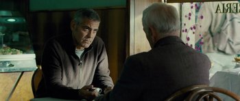 Movie still from “The American” (2010), directed by Anton Corbijn – A man sitting in front of another man holding a remote; Medium shot, Over the shoulder angle