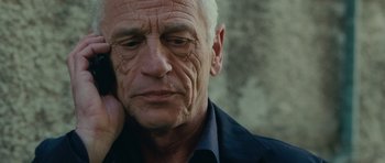 Movie still from “The American” (2010), directed by Anton Corbijn – An older man with wrinkled skin is leaning against a wall; Close Up shot, Low angle