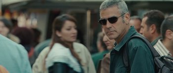 Movie still from “The American” (2010), directed by Anton Corbijn – A person wearing sunglasses on a city street; Close Up shot, Over the shoulder angle