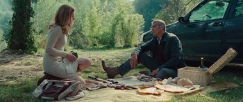 Movie still from “The American” (2010), directed by Anton Corbijn – A man and a woman sitting on a blanket in the grass; Wide shot, High angle