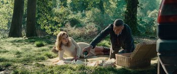 Movie still from “The American” (2010), directed by Anton Corbijn – A man and a woman are sitting on a blanket in the grass; Wide shot, High angle
