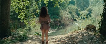 Movie still from “The American” (2010), directed by Anton Corbijn – A woman is standing on a dirt path near a river; Wide shot, Over the shoulder angle