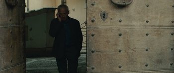 Movie still from “The American” (2010), directed by Anton Corbijn – A man standing in front of a door talking on a cell phone; Medium shot, Low angle
