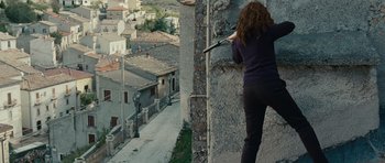 Movie still from “The American” (2010), directed by Anton Corbijn – A woman with a gun in her hand is aiming at a building; Wide shot, Low angle