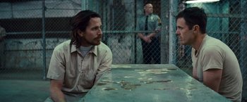 Movie still from “Out of the Furnace” (2013), directed by Scott Cooper – A man sitting at a table in front of a fence; Medium shot, Over the shoulder angle
