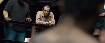 Movie still from “Out of the Furnace” (2013), directed by Scott Cooper – A man sitting at a table with his hands folded on his lap; Medium shot, Over the shoulder angle