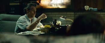 Movie still from “Out of the Furnace” (2013), directed by Scott Cooper – A man sitting in front of a tv at a table; Medium shot, Over the shoulder angle