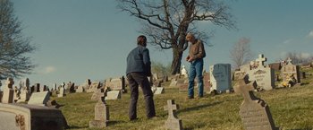 Movie still from “Out of the Furnace” (2013), directed by Scott Cooper – Two people standing in a cemetery next to a tree; Wide shot, Over the shoulder angle