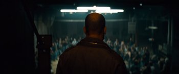 Movie still from “Out of the Furnace” (2013), directed by Scott Cooper – A man standing in front of a group of people; Medium shot, Over the shoulder angle